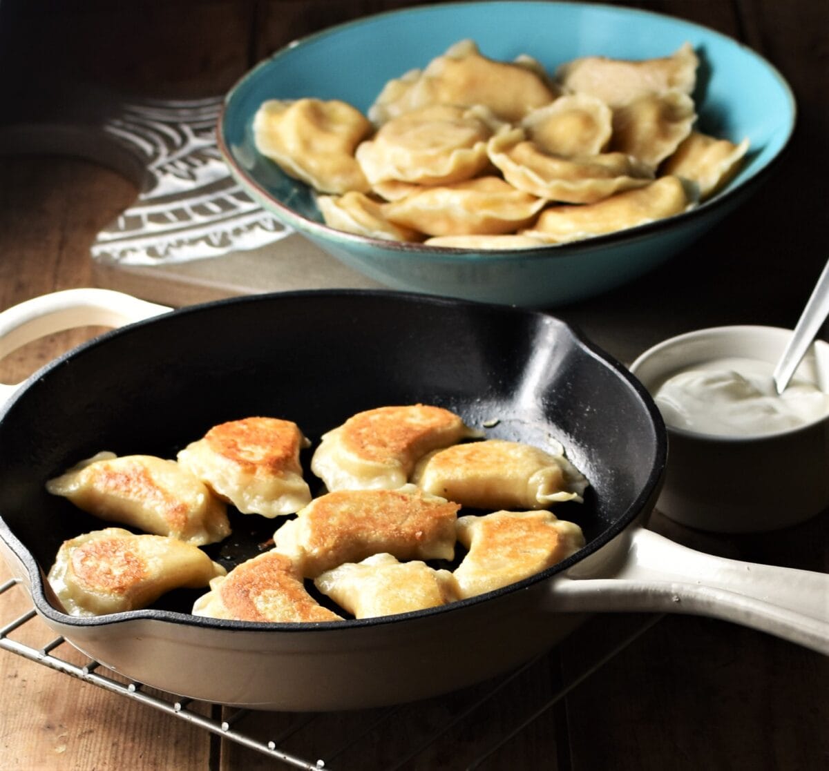 Side view of fried pierogies in pan, with perogies in blue bowl and sour cream in background.