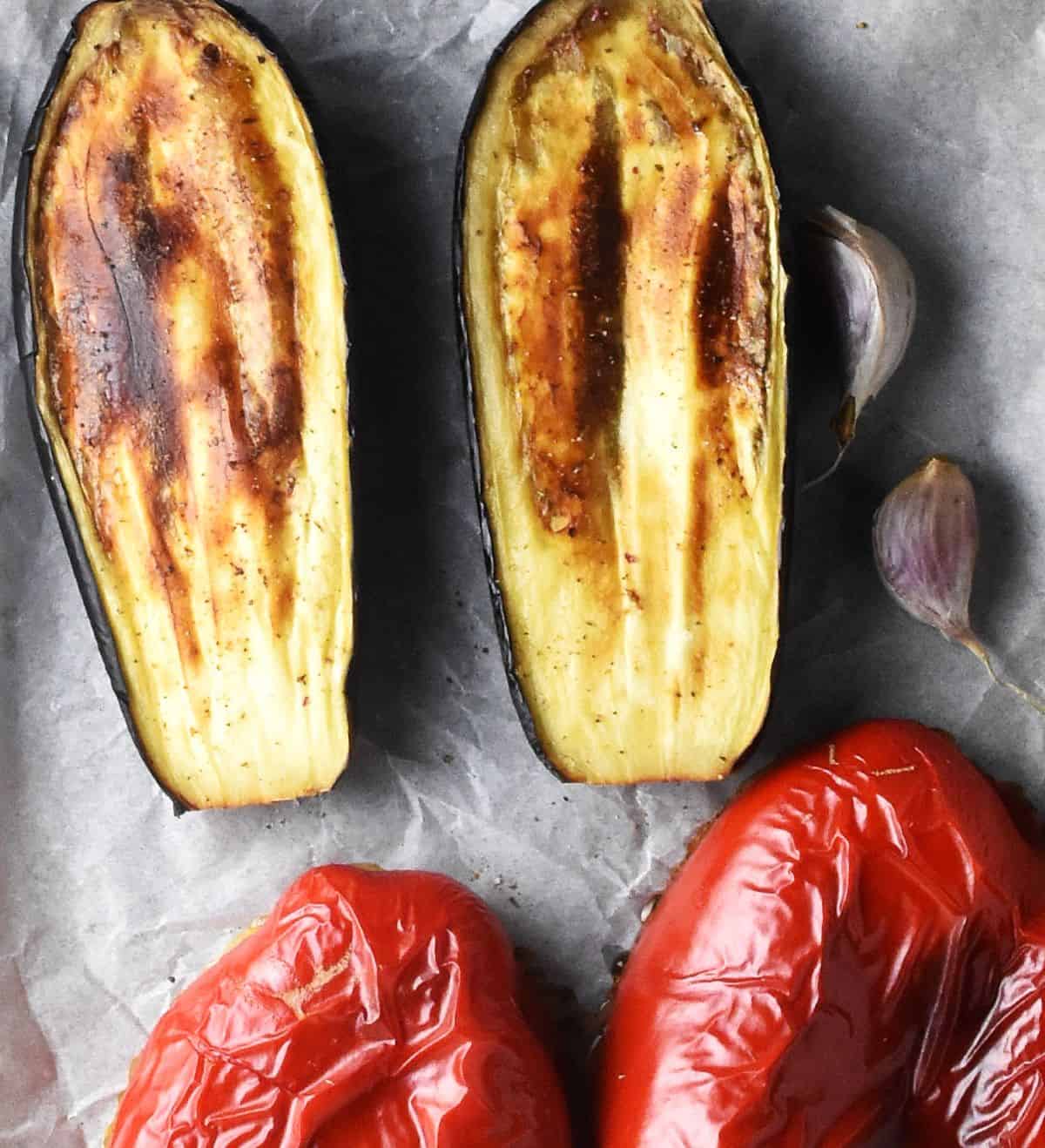 Top down view of 2 roasted eggplant halves, garlic cloves and roasted red peppers on parchment.