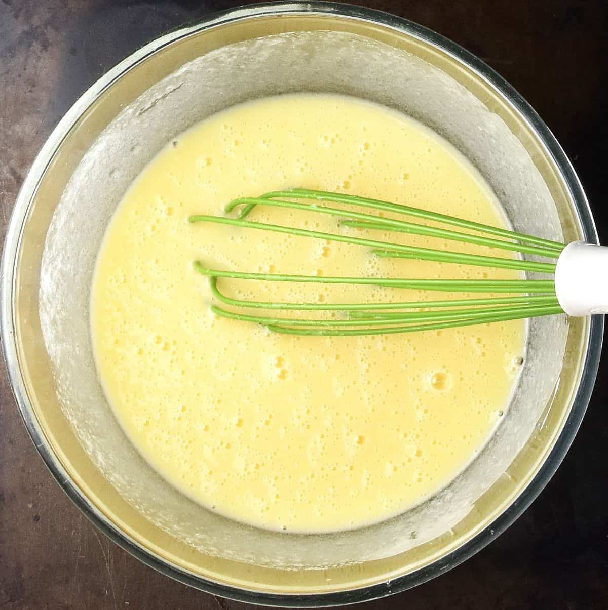 Top down view of creamy mixture in glass bowl with green whisk.