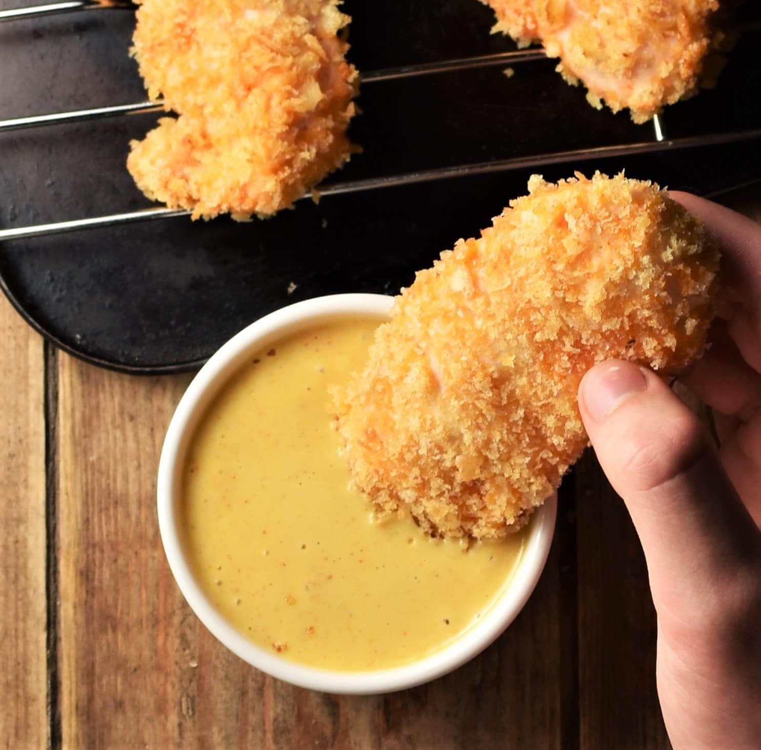 Top down view of breaded chicken tender held over mustard sauce, with chicken tenders in background.