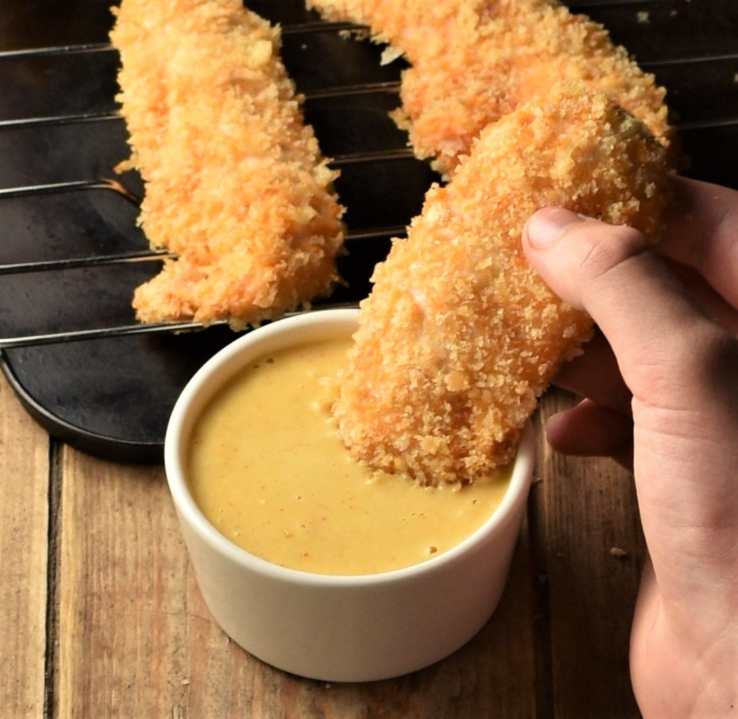 Chicken tender dipped in yellow sauce with baked tenders in background.