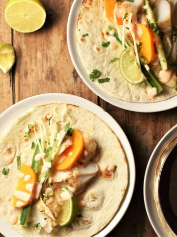 2 open fish tacos with vegetables on white plates with lime in background.