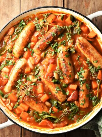 Sausage and bean casserole with vegetables and herbs in large white pan.