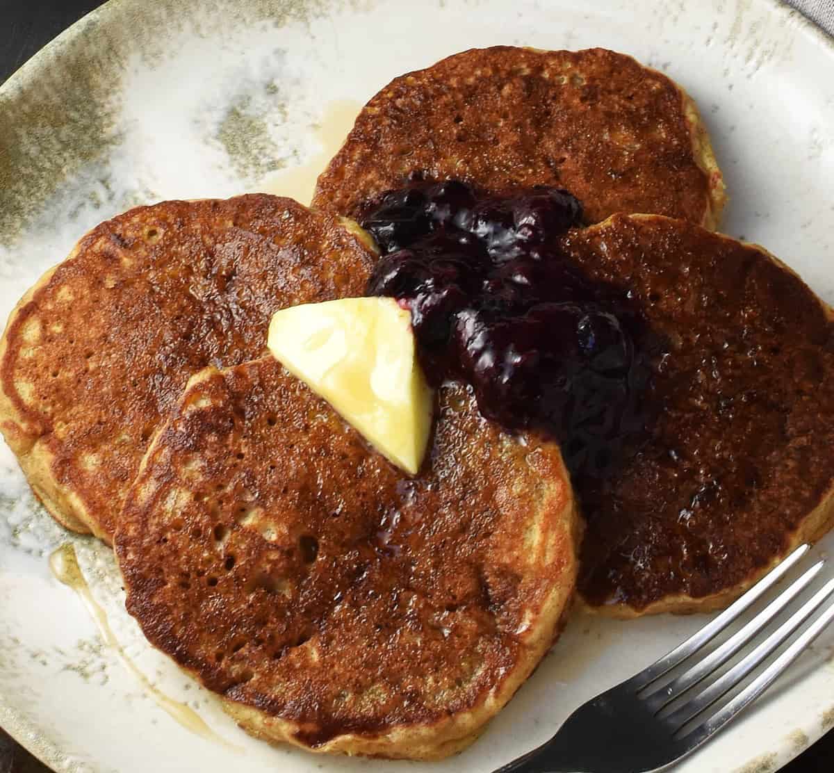 Top down close-up view of quinoa and banana pancakes with knob of butter and fruit compote on off-white plate.