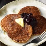 Side view of quinoa pancakes topped with butter and fruit mixture on white plate with fork.