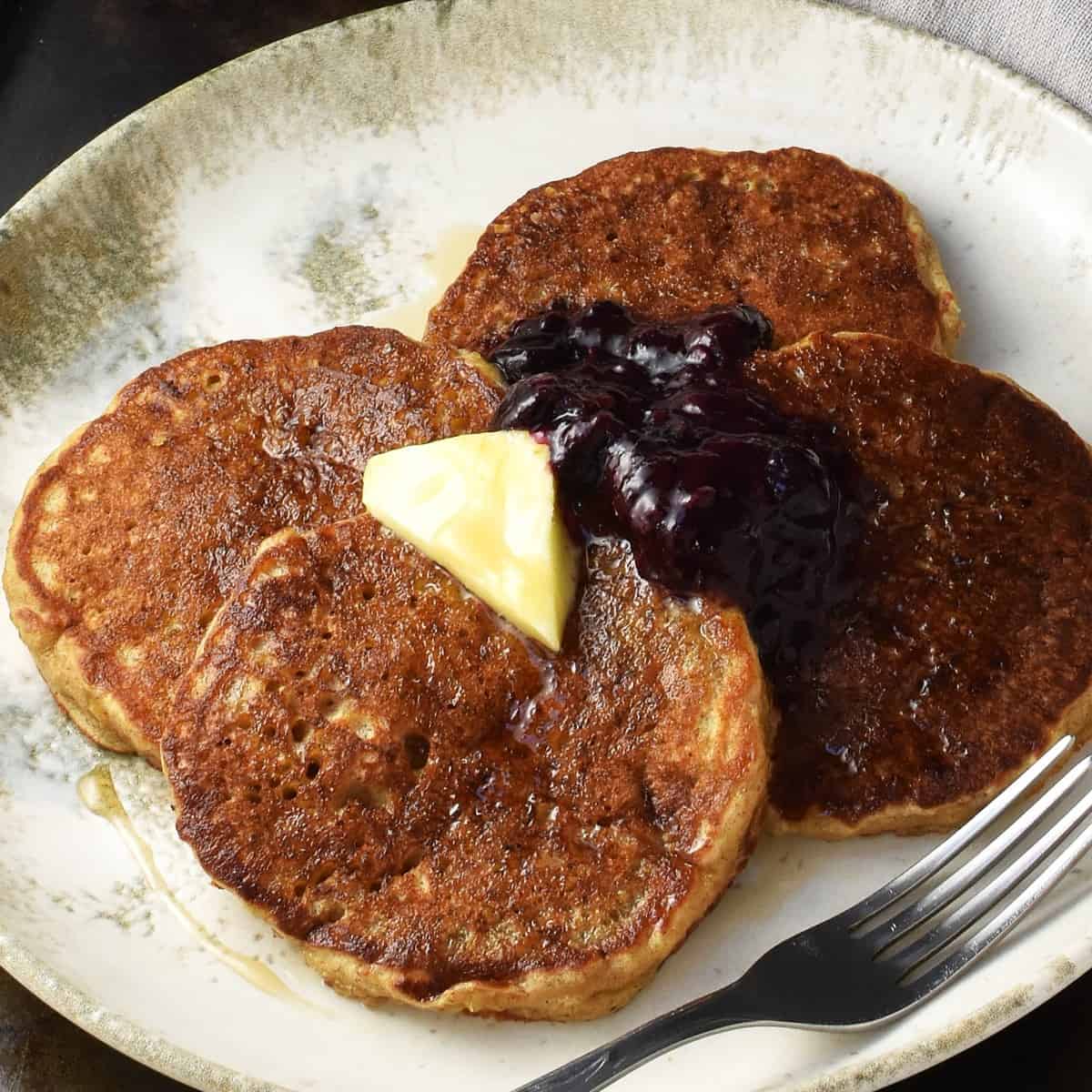 Side view of quinoa pancakes topped with butter and fruit mixture on white plate with fork.