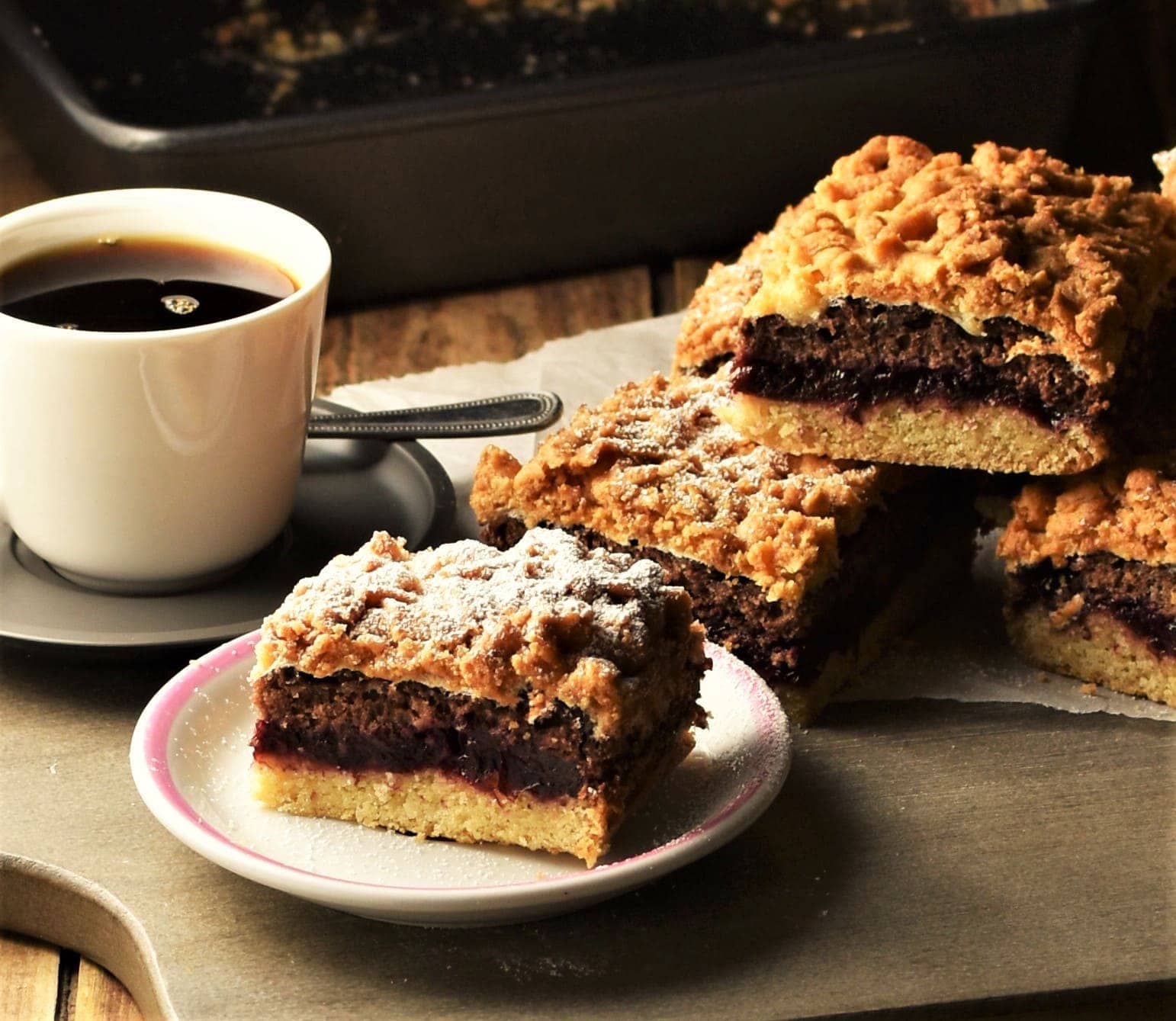 Slice of blackberry coffee cake on small plate with cake and cup of coffee in background.