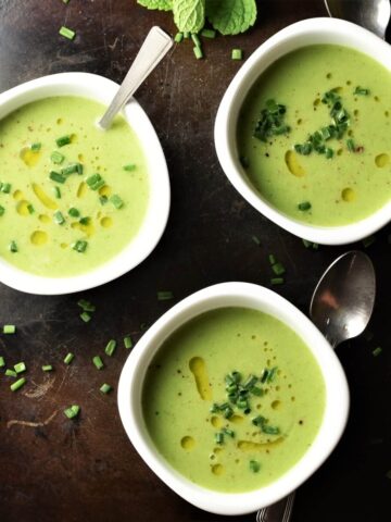 Top down view of creamy cold cucumber soup in 3 white bowls with spoons.