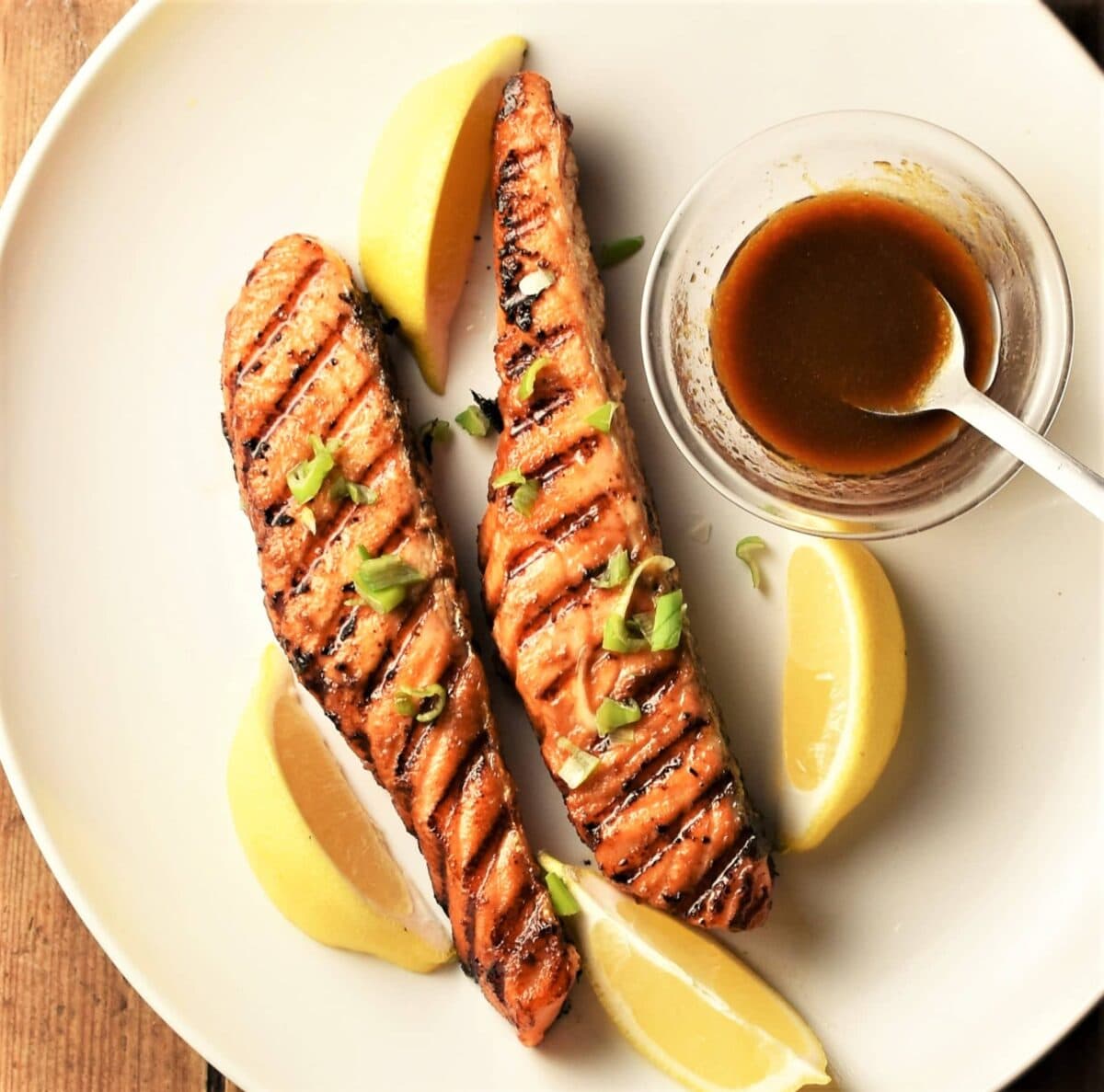 Top down view of 2 grilled salmon pieces with lemon wedges and hoisin sauce in dish.