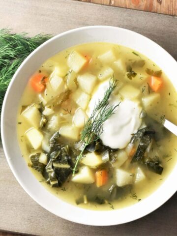 Chunky kohlrabi soup with vegetables and dollop of yogurt in white bowl with spoon and dill to the left.