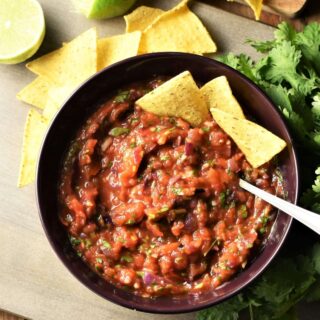Roasted tomato salsa in bowl with nachos and spoon, nachos, limes and herbs in background.