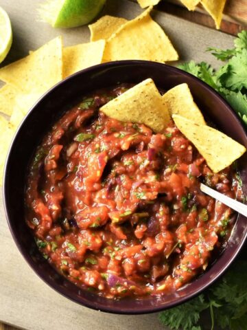 Roasted tomato salsa in bowl with nachos and spoon, nachos, limes and herbs in background.