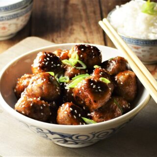Close-up view of meatballs with glaze in bowl with chopsticks and rice in background.
