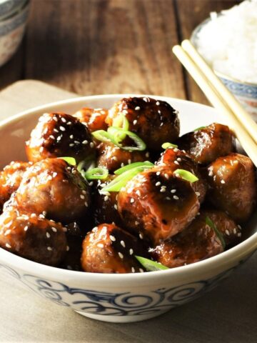 Close-up view of meatballs with glaze in bowl with chopsticks and rice in background.