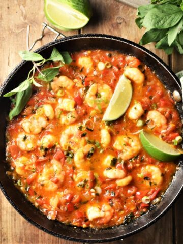 Shrimp in tomato coconut sauce with lime wedges and herbs in pan.