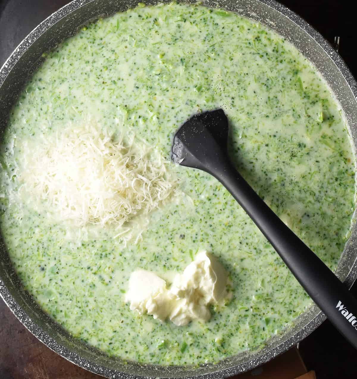 Top down view of creamy broccoli sauce with grated parmesan and cream cheese in large pan with black spatula.