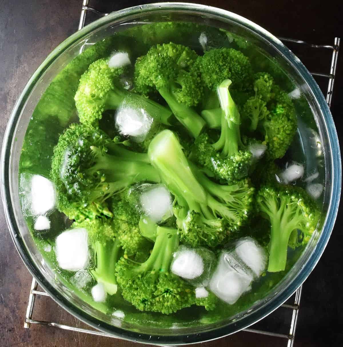 Broccoli florets in water with ice cubes in glass bowl.