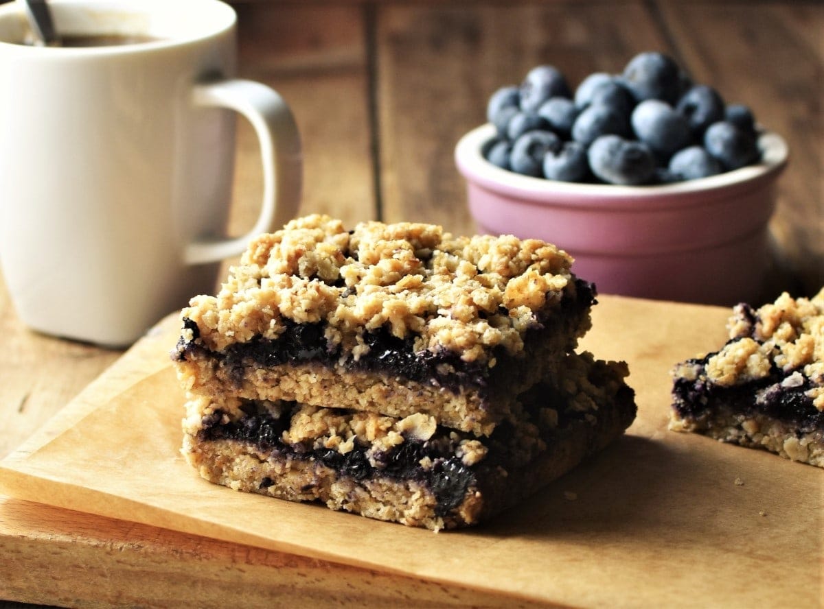 Side view of blueberry oatmeal bars on top of board, with white cup and blueberries in background.