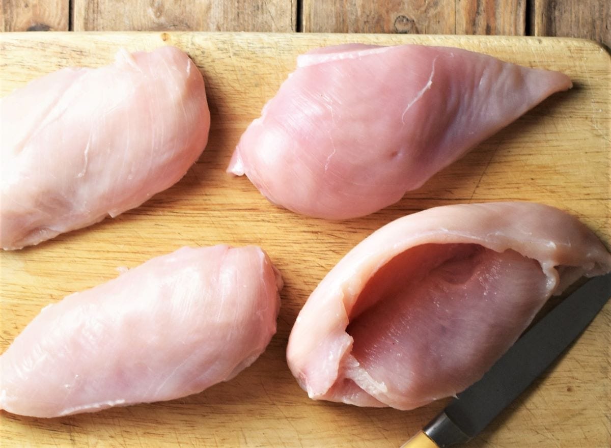 4 raw skinless chicken breasts on top of cutting board with knife.