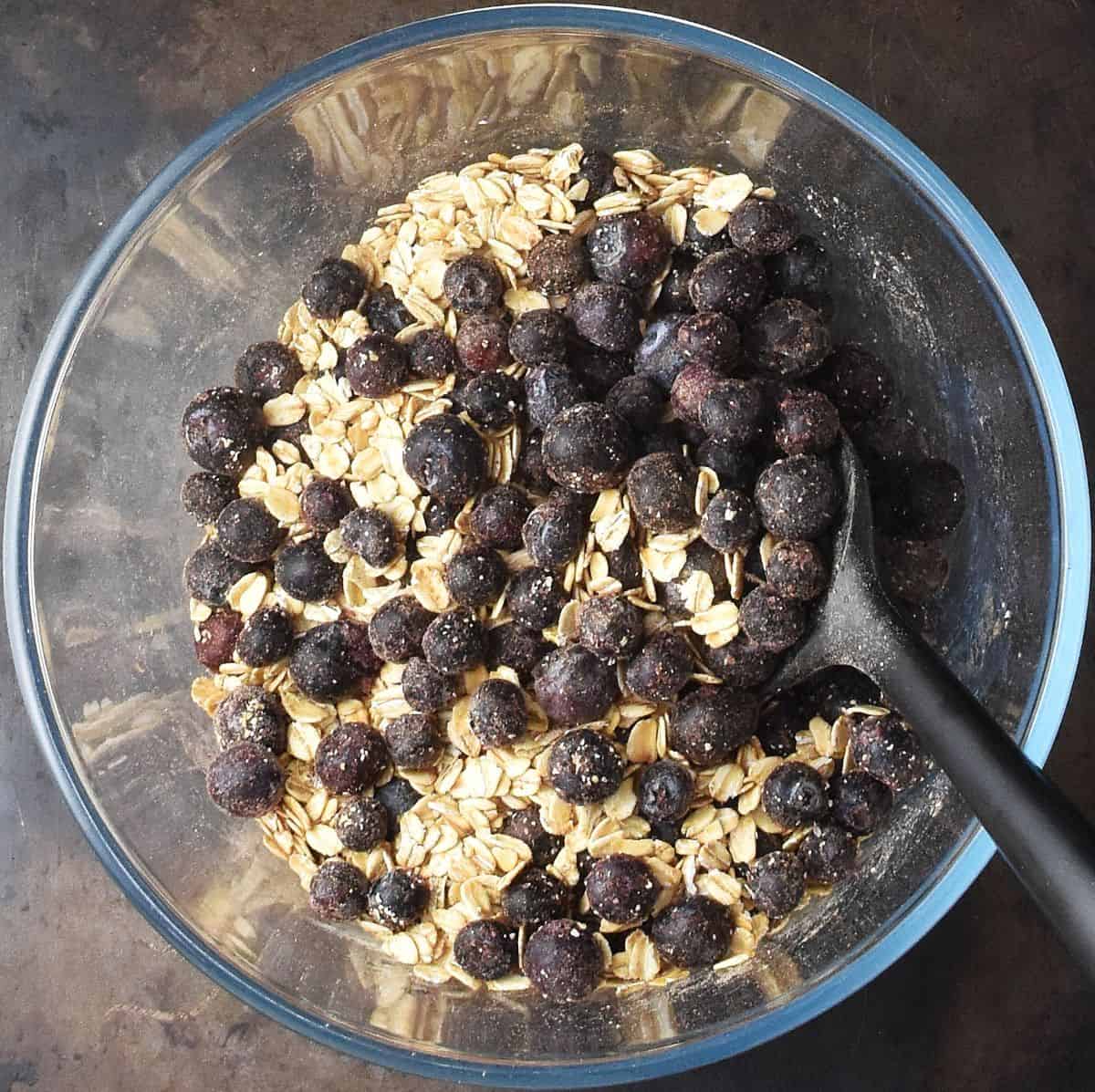 Top down view of blueberries with oat mixture in glass bowl and black spatula.