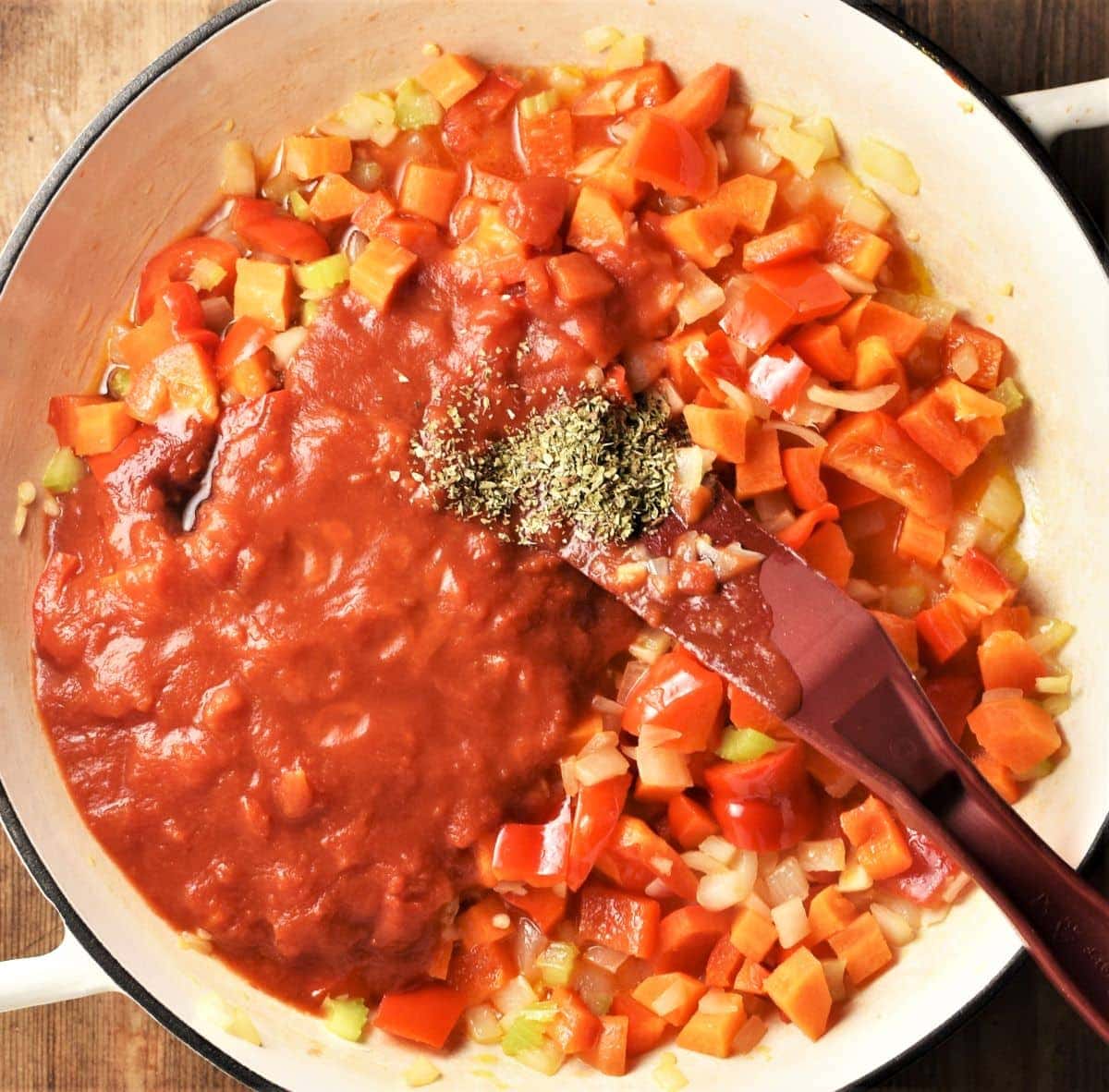 Chopped vegetables and tomato puree in white shallow pan with red spatula.