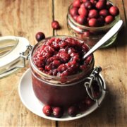 Side view of cranberry chutney in open jar with spoon and cranberries in background.