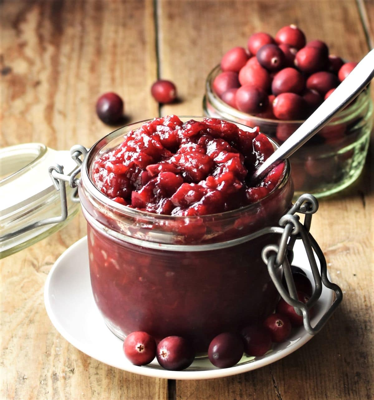 Side view of cranberry chutney in open jar with spoon on top of white plate with fresh cranberries in background.