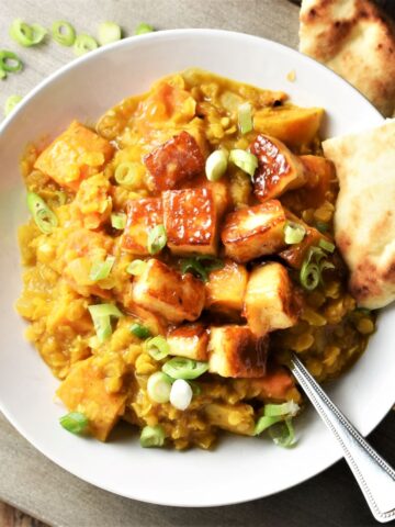 Top down view of sweet potato dahl with fried halloumi cubes in white bowl with naan and spoon.