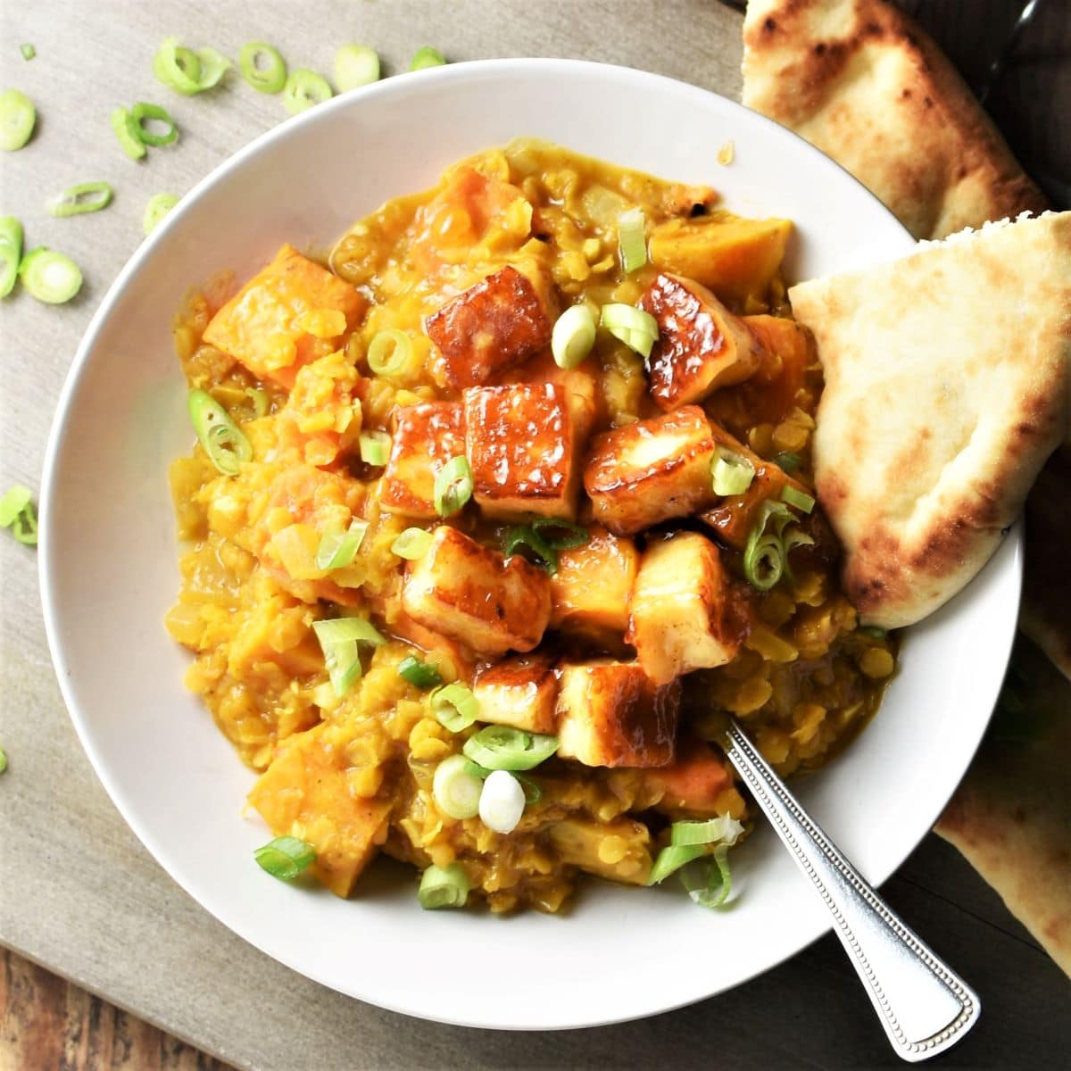 Top down view of sweet potato dhal with fried halloumi cubes in white bowl with naan and spoon.