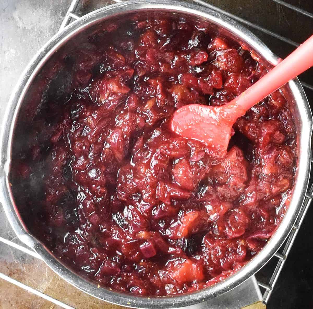 Cooked cranberry chutney in pot with red spatula on top of rack.
