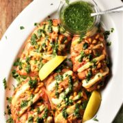 Baked hasselback chicken breasts with halloumi, herb sauce and lemon wedges in white dish.