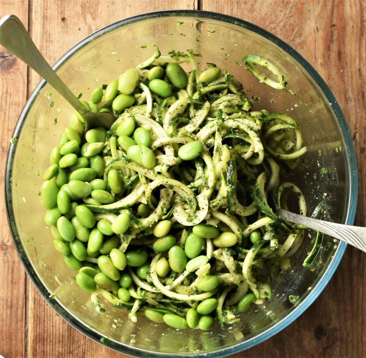 Zoodles with pesto and edamame beans in mixing bowl with 2 forks.