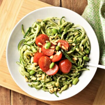 Pesto zoodles salad with cherry tomatoes and beans in white bowl with fork.
