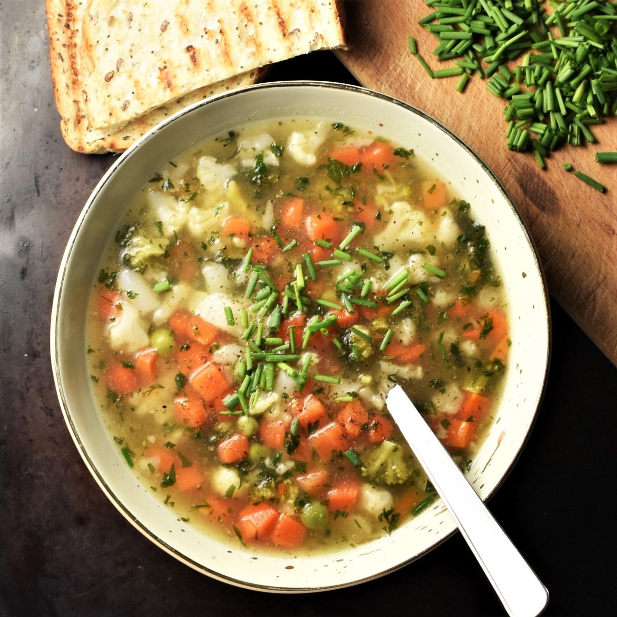 Chunky vegetable soup in green bowl with spoon, fresh chives and grilled bread in background.