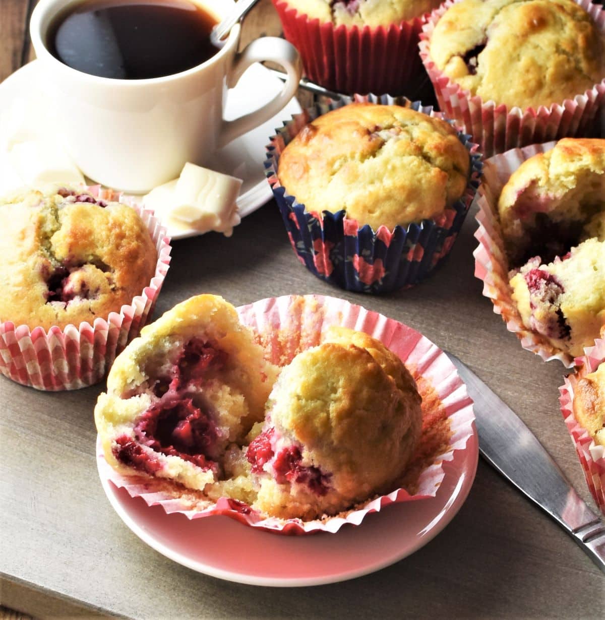 Side view of raspberry muffins in paper cases and coffee in background.