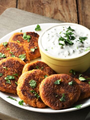 Side view of sweet potato fritters on top of plate with yogurt dip in yellow dish.