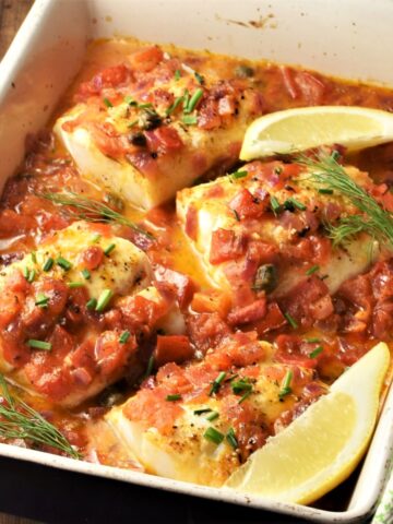 Side view of 4 cod pieces in tomato sauce in casserole dish with lemon wedges.