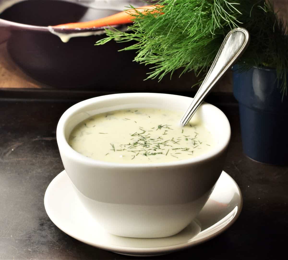 Side view of creamy dill sauce in white bowl with spoon and fresh dill in background.