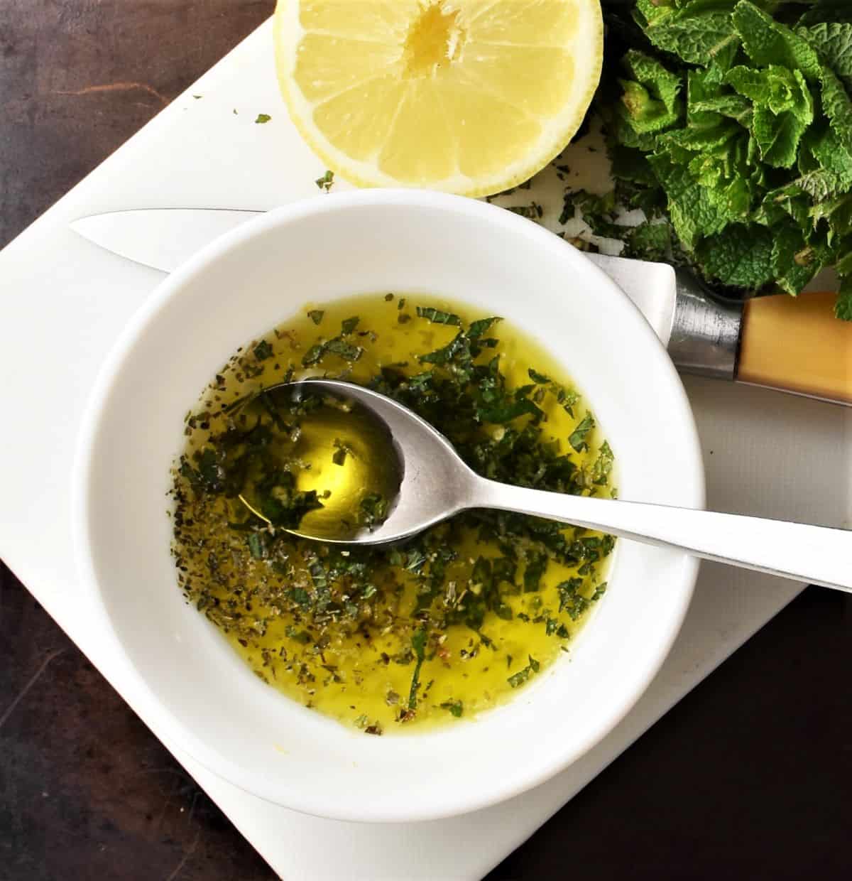 Salad dressing in white bowl with spoon, lemon and herbs.