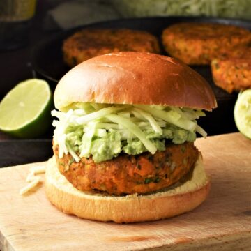 Side view of sweet potato burger with cabbage on top of wooden board with lime and burgers in background.