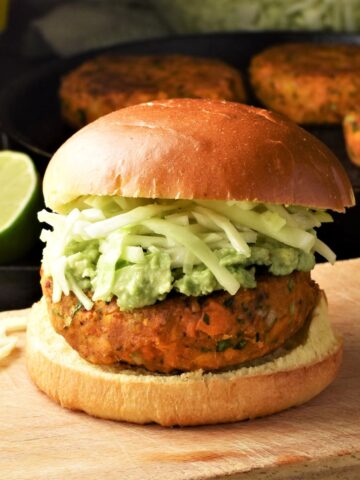 Side view of sweet potato burger with cabbage on top of wooden board with lime and burgers in background.