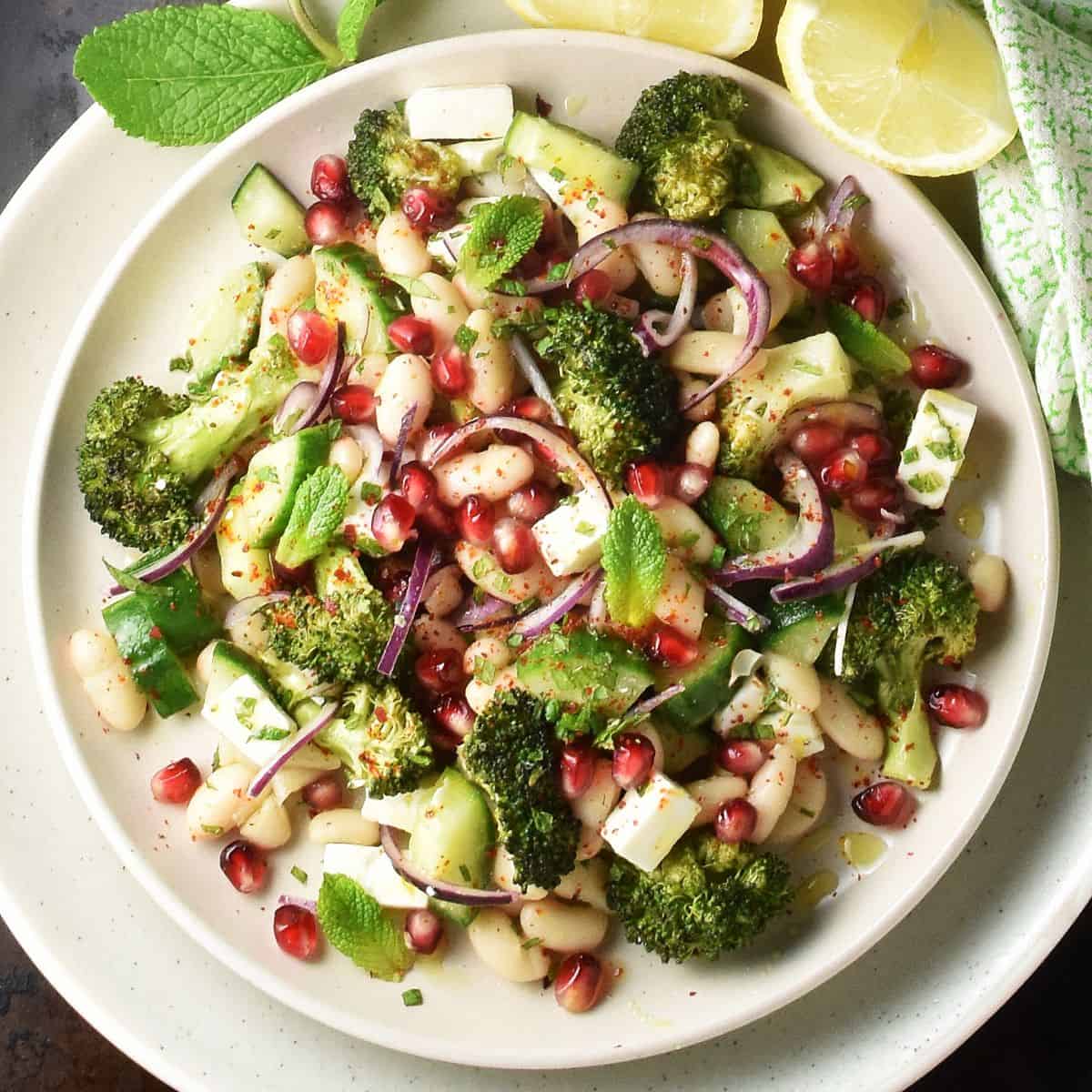 Broccoli salad with beans, feta and pomegranate on white plate with mint and lemon in background.