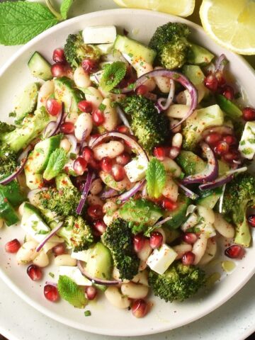 Top down view of roasted broccoli and bean salad with feta and pomegranate seeds on white plate with lemon wedges.