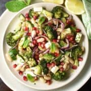 Top down view of roasted broccoli and bean salad with feta and pomegranate seeds on white plate with lemon wedges.