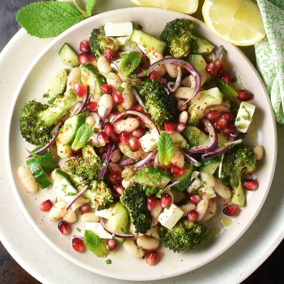 Top down view of roasted broccoli and bean salad with feta and pomegranate seeds on white plate with lemon wedges.