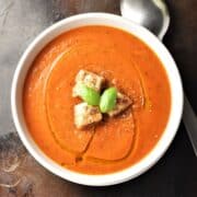 Top down view of creamy tomato zucchini soup with croutons and spoon in background.