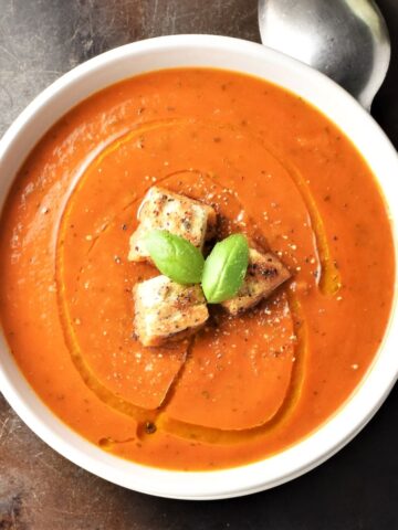 Top down view of creamy tomato zucchini soup with croutons and spoon in background.