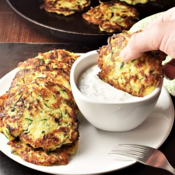 Side view of potato zucchini fritters on top of white plate with fritter dipped in yogurt dip.