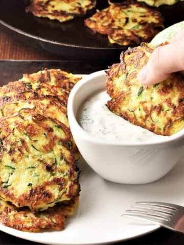 Side view of potato zucchini fritters on top of white plate with fritter dipped in yogurt dip.