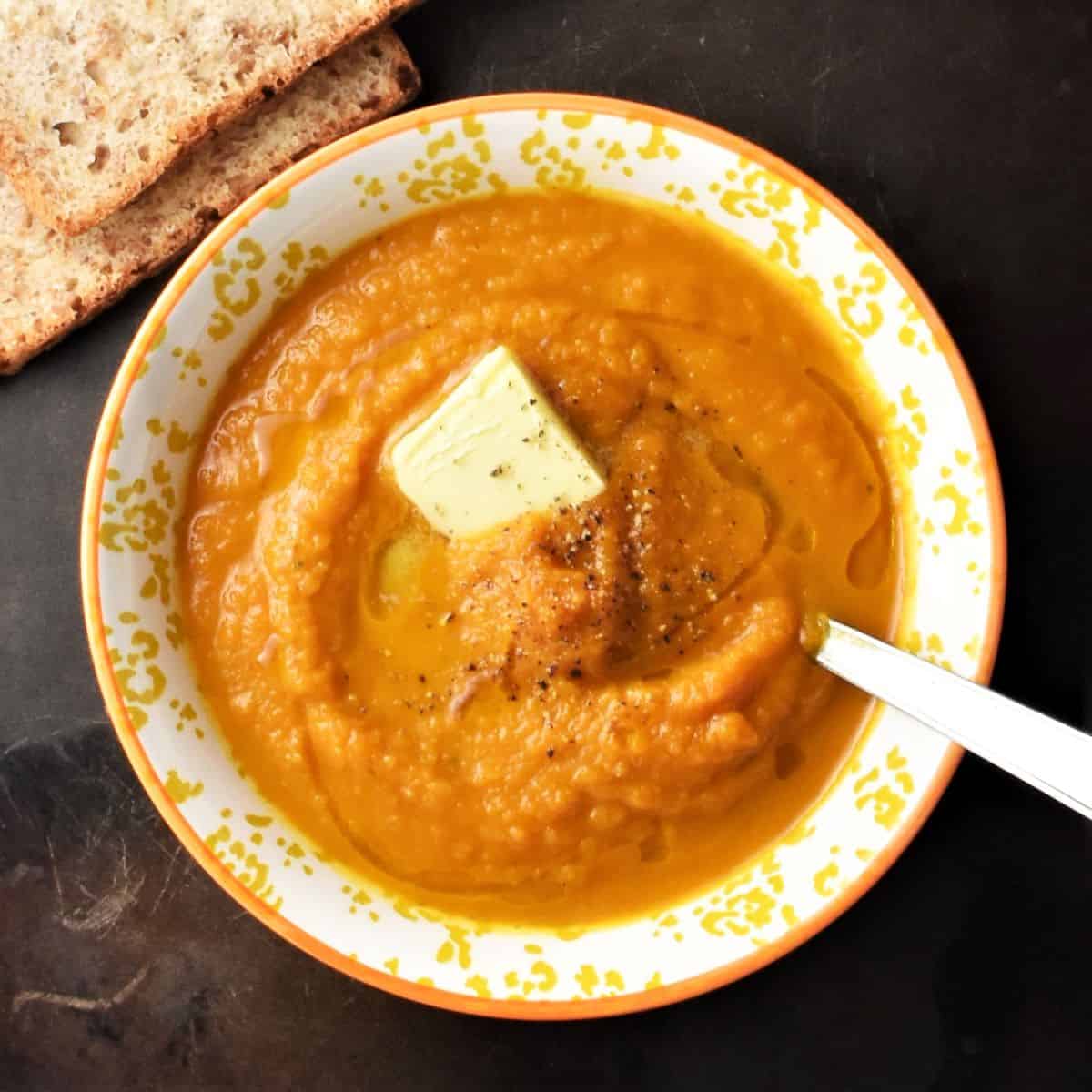 Top down view of creamy pumpkin carrot soup with butter in yellow bowl with spoon.